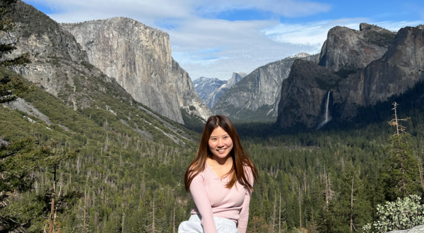 Female, early 20s, with dark hair, sitting on stone ledge in front of trees and mountain at Yosemite National Park