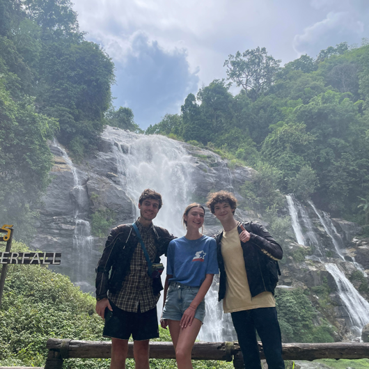 three people in their 20s, one female standing between two males, with a waterfall and green trees behind them