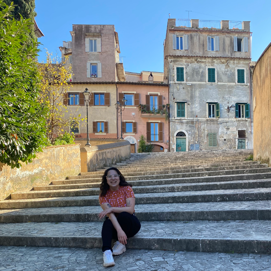 Female, early 20s, with dark hair, sitting on stone steps in front of old stone buildings in Italy