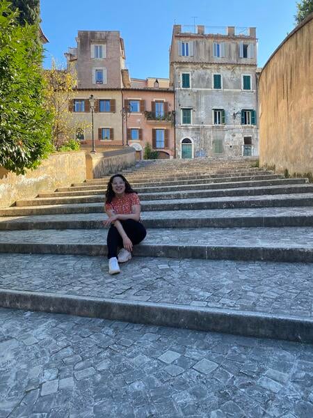 Female, early 20s, with dark hair, sitting on stone steps in front of old stone buildings in Italy