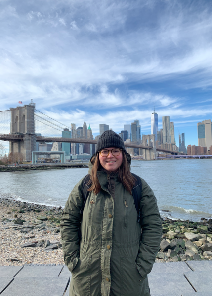 Female, early 20s, with brown hair and wearing glasses, standing near water, with the Brooklyn Bridge and the New York City Skyline behind