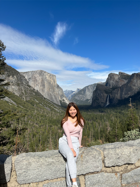 Female, early 20s, with dark hair, sitting on stone ledge in front of trees and mountain at Yosemite National Park